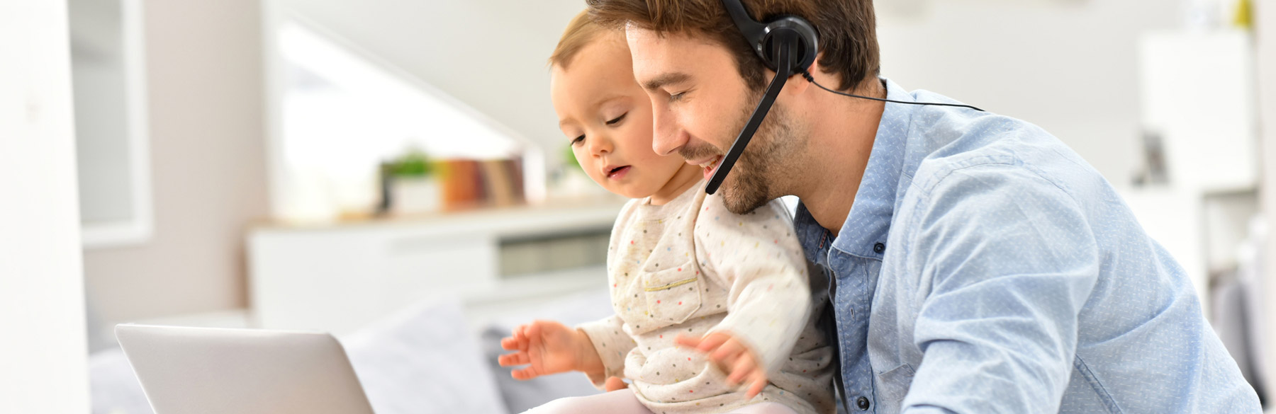 Ein Vater arbeitet mit einem Headset an einem Laptop, während sein Kind neben ihm sitzt.